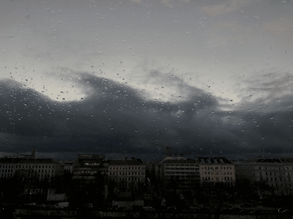 Wir schauen durch eine mit Regentropfen gesprenkelte Fensterscheibe auf das trübe Landschaftsbild einer Stadt.